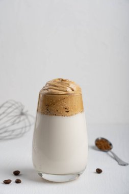 Tek bardak popüler Kore dalgona içeceği hazır kahve tozu, şeker ve sıcak su kırbaçlanarak yapılır ve beyaz ahşap zemin üzerine soğuk süt ve mutfak gereçleri eklenir. Ses yönelimi