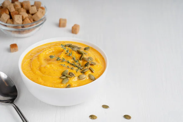 stock image Vegetarian homemade organic pumpkin soup made of pureed vegetables decorated with seeds and fresh thyme served in bowl with spoon and croutons on white wooden background. Image with copy space