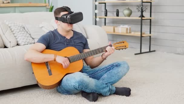 Young man learning to play guitar using VR 360 headset while sitting on carpet at home — Stockvideo