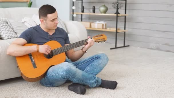 Молодий чоловік грає на акустичній гітарі, сидячи на килимі у вітальні — стокове відео