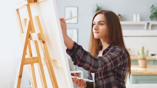 Une jeune femme peintre dessine une toile sur un chevalet à la maison — Video