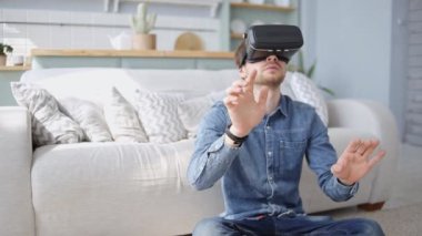 Genç adam evde sanal gerçeklik gözlüğü kullanıyor. VR kulaklıklı bir erkek halının üzerinde oturuyor ve dijital simülasyonda nesnelere dokunuyor. Gerçekçi 3D görselleştirme, yenilikçi teknoloji