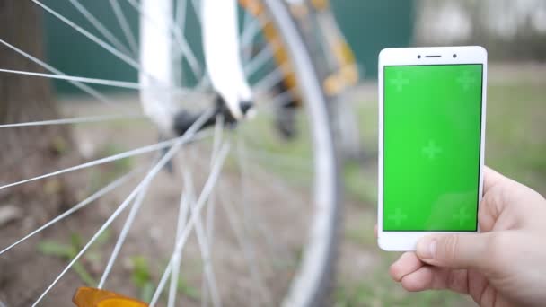 Homem mão segurando telefone inteligente móvel com tela verde mock-up, bicicleta no fundo — Vídeo de Stock