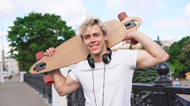 Glad blond man med hörlurar som håller longboard bakom huvudet och leende — Stockvideo