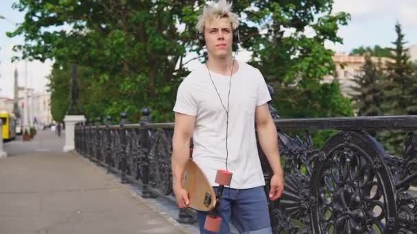 Joven hombre rubio sosteniendo longboard deck en las manos y caminando en un día de verano — Vídeos de Stock