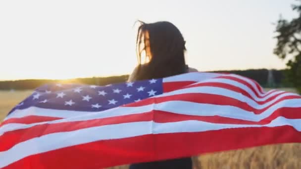 Joven chica sonriente corriendo con la bandera de EE.UU. sobre el campo de trigo y se da la vuelta en cámara lenta — Vídeos de Stock