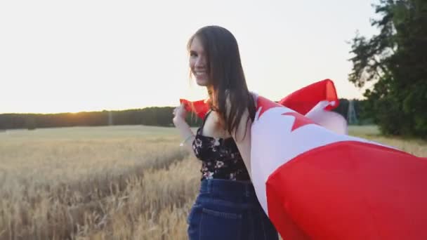 Молодая улыбающаяся девушка бежит с флагом Канады по пшеничному полю и разворачивается в замедленной съемке — стоковое видео