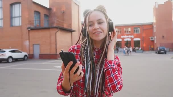 Chica adolescente de moda con rastas escuchando música en los auriculares y utilizando el teléfono inteligente — Vídeos de Stock