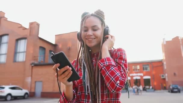 Chica adolescente de moda con rastas escuchando música en los auriculares y utilizando el teléfono inteligente — Vídeos de Stock