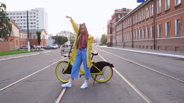 Menina com dreadlocks e capa de chuva amarela posando com bicicleta amarela vintage, sorrindo e se divertindo — Vídeo de Stock