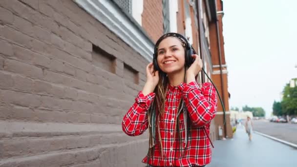 Elegante chica adolescente con rastas escuchando música en los auriculares y caminando por la calle — Vídeo de stock
