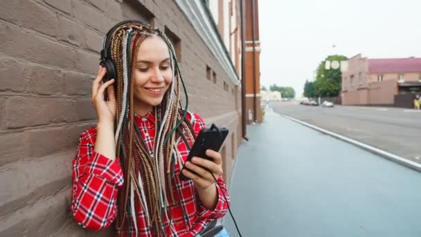 Beautiful smiling girl with dreadlocks and headphones enjoying listens to music in city — Stock Video