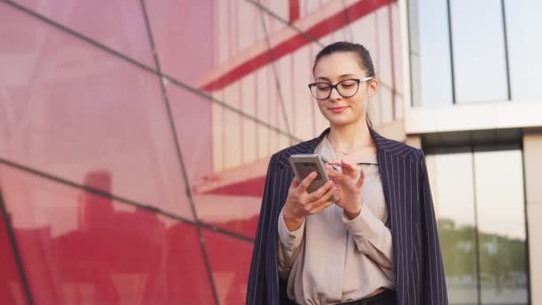 Retrato de mujer de negocios sonriente con gafas usando un teléfono inteligente, edificio moderno en el fondo — Vídeos de Stock