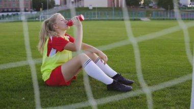 Genç futbolcu kız antrenmandan sonra dinleniyor çimenlerde oturuyor ve şişeden su içiyor..