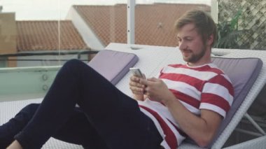 Genç adam bir yaz günü güverte sandalyesinde akıllı telefon kullanıyor. Tropik bir otelin çatısında dinlenen bir adam..
