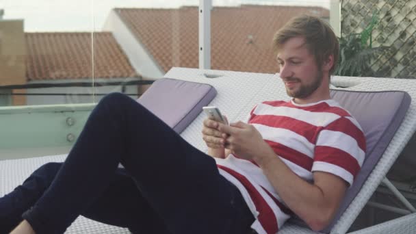 Jeune homme utilisant un téléphone intelligent couché sur une chaise longue un jour d'été. Homme relaxant sur le toit d'une station tropicale . — Video