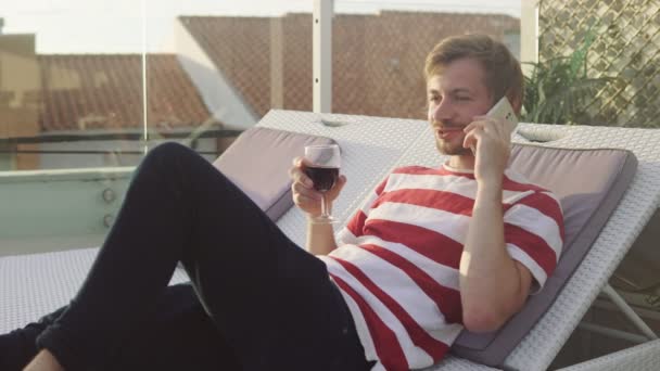 Retrato del hombre relajado sosteniendo una copa de vino y hablando por teléfono inteligente en una azotea de un resort de lujo . — Vídeos de Stock