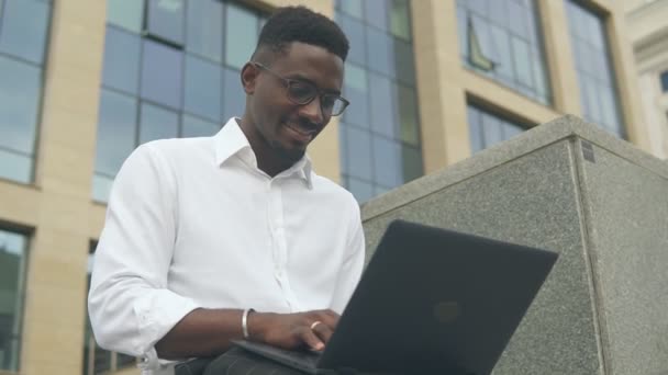 Young african american businessman working on laptop outdoors on a background of corporate building. — Stock Video