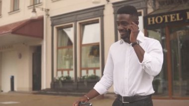 Afro-Amerikan turistin tatilde, elinde bir bavulla otelin dışında, akıllı telefondan konuşan, ulaşımı bekleyen bir çanta..