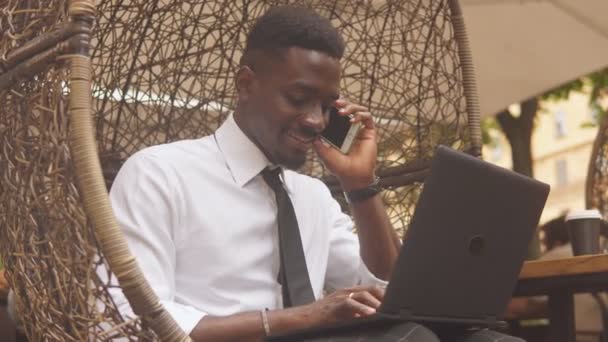 Close up of young african american businessman in white shirt working with laptop outdoors talking on smartphone. Slow motion — Stock Video