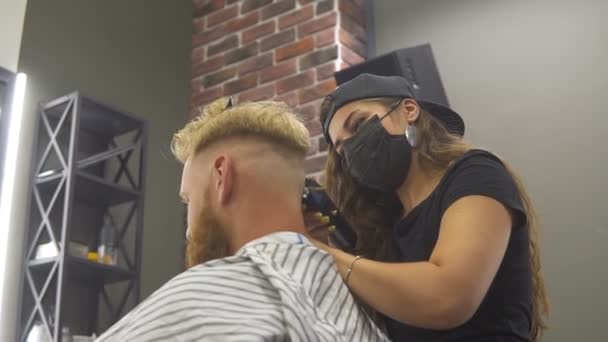 Homem caucasiano jovem recebendo novo corte de cabelo na barbearia moderna. Profissional jovem barbeiro menina na máscara fazendo corte de cabelo para seu cliente . — Vídeo de Stock