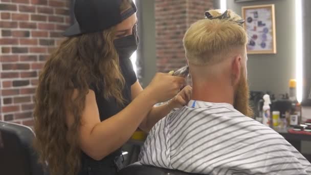 Jovem barbeiro profissional em máscara fazendo corte de cabelo para seu cliente. Fechar — Vídeo de Stock