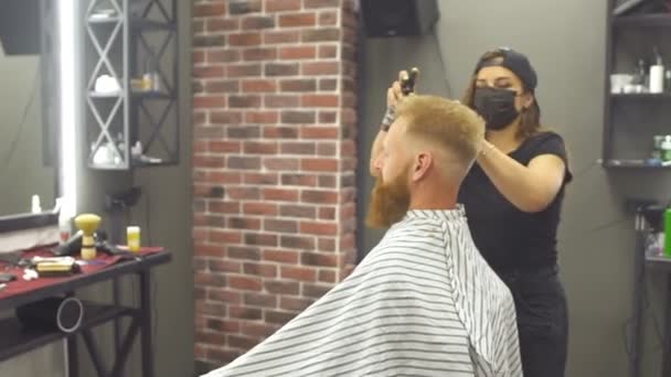 Young ginger man getting new haircut in the modern barbershop. Professional young barber girl in mask doing haircut for her client.