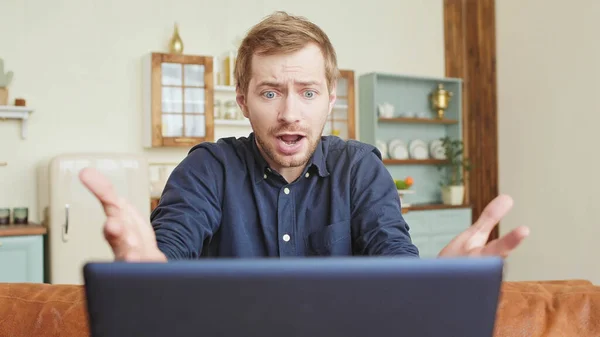 Молодой человек в напряженной рубашке работает за столом в гостиной, кричит на экран ноутбука и злится Лицензионные Стоковые Фото