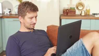 Gülümseyen adam boş vakitlerini kanepede geçiriyor ve modern evde dizüstü bilgisayar kullanıyor..