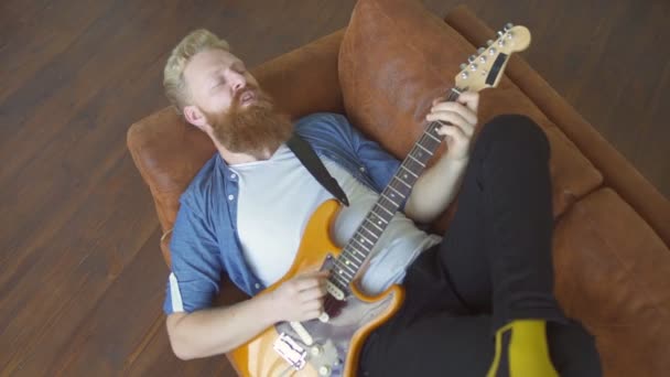 Happy baardman hipster spelen van de elektrische gitaar terwijl liggend op de bank — Stockvideo