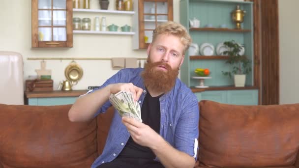 Pria berambut merah makan uangnya duduk di sofa di rumah. — Stok Video