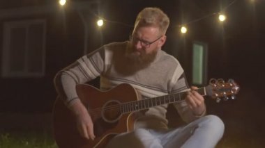 Yürüyüşte kamp ateşinin yanında akustik gitar çalan sakallı genç bir adam.