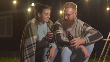 Mutlu genç kadın ve adam şenlik ateşinde dinlenirken bira şişelerini tokuşturup birbirleriyle konuşuyorlar.