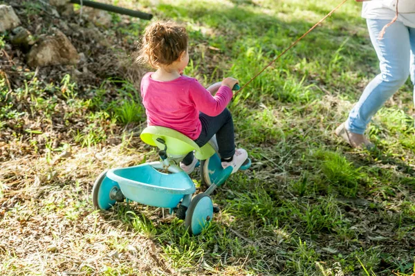 toddler girl in nature sitting on her velotrol being pulled by a rope. she has her back to the camera