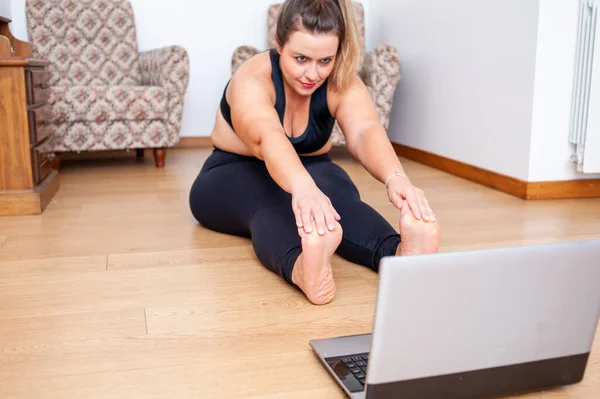 plus size woman streching her body on a  gym class at home. She is using her computer to watch video lessons.