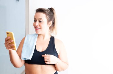 Egzersiz yaptıktan sonra cep telefonuna bakan mutlu bir kadın. Omzunda mavi bir havlu var. Boşluğu kopyala.