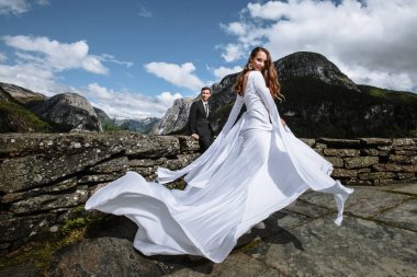 Dağların arka planında yeni evliler. Gelin, damadın arkasında, uzun bir gelinlik içinde.