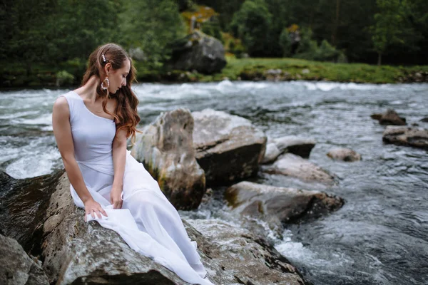 A girl in underwear and a light transparent Cape sits on a rock by the river