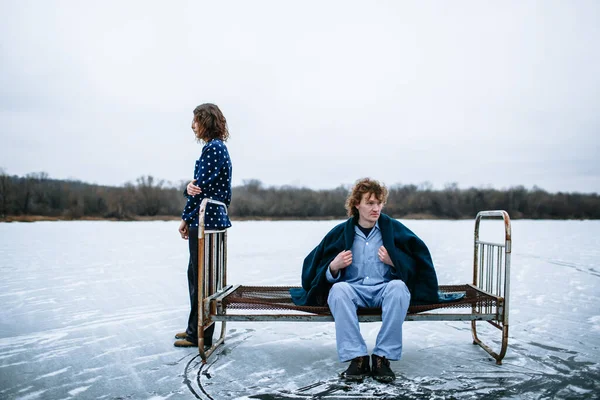 A guy and a girl in pajamas on the ice of a frozen lake. The guy is sitting on an iron bed, the girl next to him.