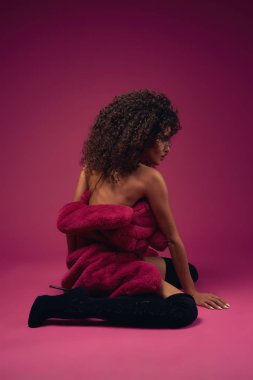 Parlak bir arka planda oturan bir kürk ve botlar yarım çıplak model