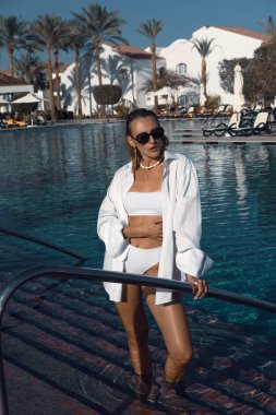 Havuz kenarında beyaz mayo ve gömlekle poz veren genç kız.