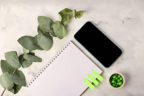 Blank notebook with push pins near it on the marble background.Mobile telephone with black screen..Fresh eucalyptus branch on the back.Copy space for text.Office mockup concept.