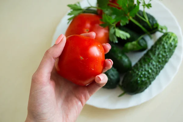 A tomato on the woman hand. A fresh tomatoes, sliced cucumbers and sprig of parsley on the white plate on the light table. Seasoning vegetables on a table. Healthy ingredient for salad.