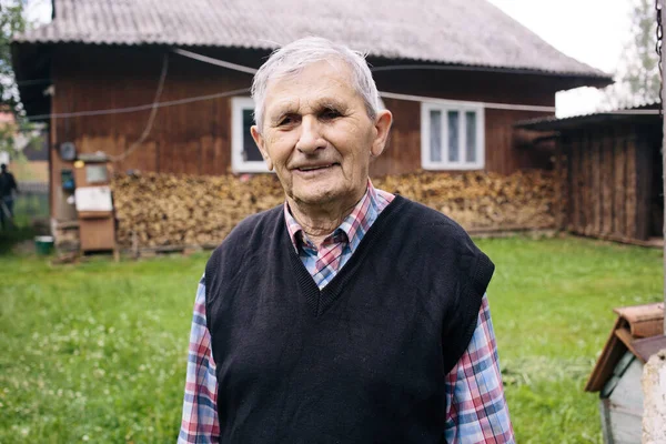 A Happy old grandfather smiling and standing on the background of a house and wood in the yard in the village. The older generation.