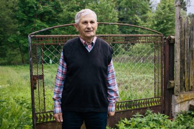 Köyün bahçesindeki bahçe çitlerinin arkasında gülümseyen ve duran mutlu yaşlı bir büyükbaba. Eski nesil..