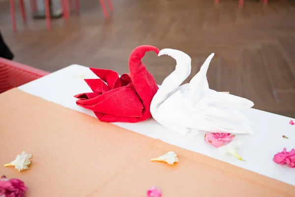 Red and white folded swan bird shape napkin in a restaurant on a white table at a resort. A flower petals and glasses on the table. Romantic dinner for newlyweds. Sunset.