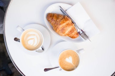 Bir fincan çay tabağında beyaz bardakta iki kahve ve beyaz tabakta bir kruvasan, bir çatal ve bir bıçak bir kafede bir yazlık terasta. Arka planda telefonu olan bir kız. Üst Manzara. düz yat