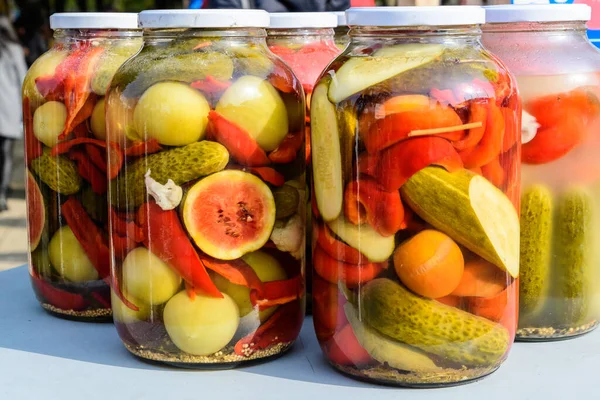 Five glass jars with mixed pickles on a white table for sale at a food market with pickled cucumbers, green tomatoes, red peppers, melon, cauliflower, side view with soft focus of tasty healthy food