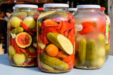 Salatalık turşusu, yeşil domates, kırmızı biber, kavun ve karnabaharın olduğu bir gıda pazarında satılmak üzere beyaz bir masada karışık turşulu üç kavanoz.