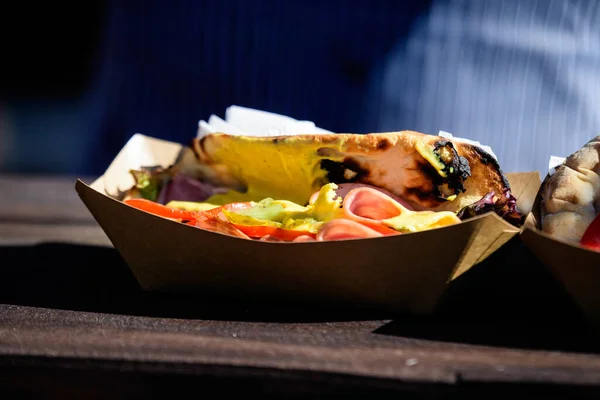 One fresh healthy Brazilian sandwich or burger with ham, green salad, pickled cucumbers and sauce is ready to eat in a brown box on a table at a street food market festival, side with soft focus